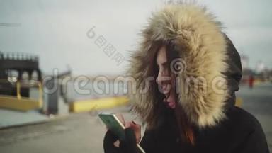 使用智能手机技术应用程序的美女穿着一件带绒毛罩的<strong>保暖</strong>夹克在街上行走的特写镜头